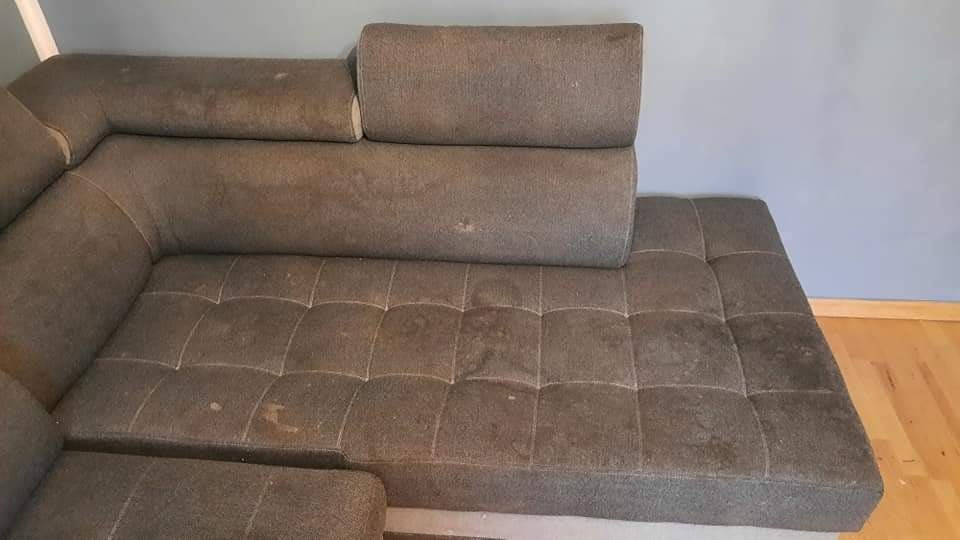 Szary narożnik z wyraźnymi plamami i zabrudzeniami na tapicerce, widoczne ślady użytkowania na siedzisku i oparciu.