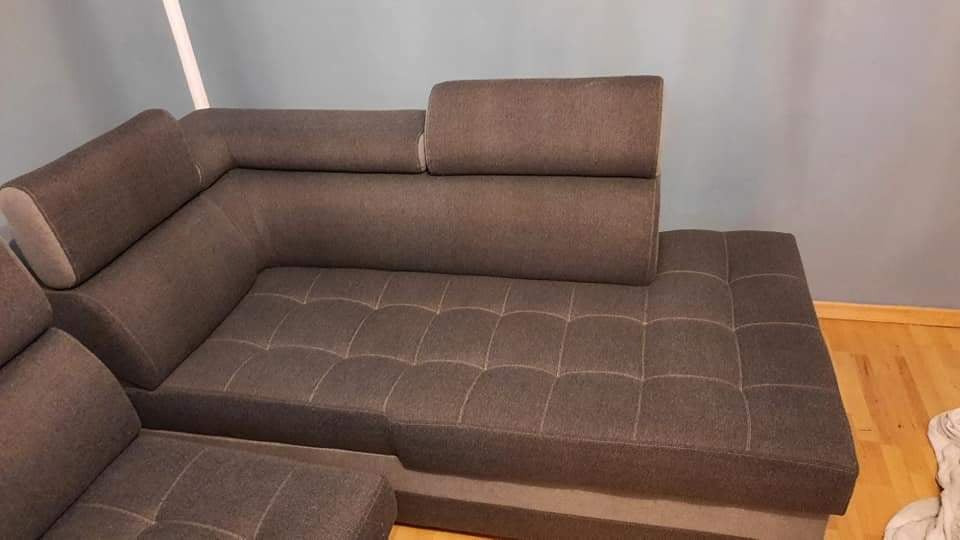 Szara, modułowa kanapa narożna z regulowanymi zagłówkami i pikowanym siedziskiem, sfotografowana w pomieszczeniu o jasnych ścianach i drewnianej podłodze.