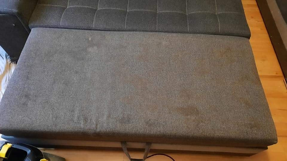 Szary narożnik z widocznymi plamami na tapicerce siedziska, częściowo rozłożony, z fragmentem odkurzacza piorącego w lewym dolnym rogu.