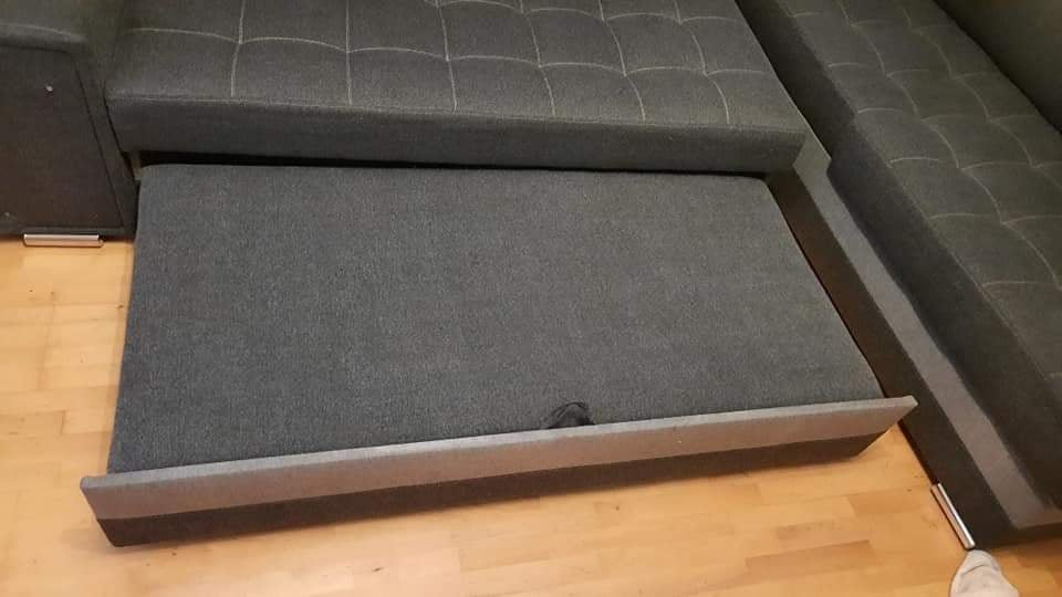 Wysunięta szuflada z szarej tapicerki z narożnika modułowego na drewnianej podłodze, widoczny fragment siedziska z pikowaniem.