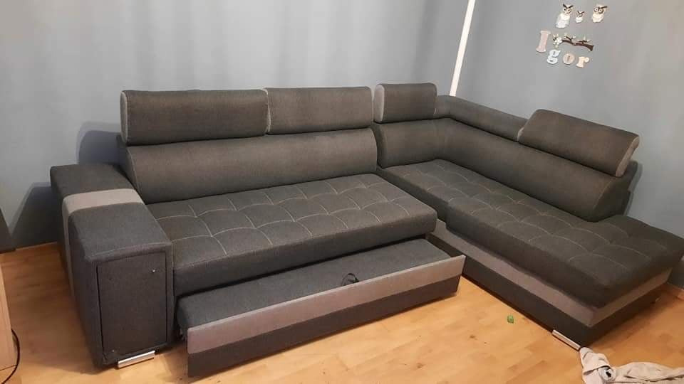 Rozłożona narożna kanapa w odcieniach szarości z wysuwanym pojemnikiem na pościel, widoczne drewniane panele podłogowe oraz dekoracja ścienna z imieniem Igor.