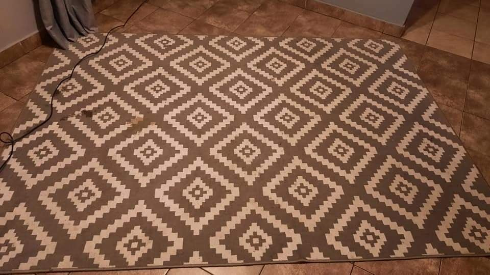 Szaro-biały dywan w geometryczne wzory leży na brązowej podłodze w mieszkaniu w Rudzie Śląskiej. Widoczny kabel od odkurzacza.