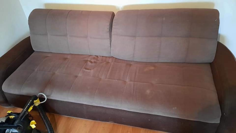 Brązowa, dwuosobowa kanapa z widocznymi plamami, częściowo wyczyszczona, obok odkurzacz piorący z żółtymi elementami na drewnianej podłodze.