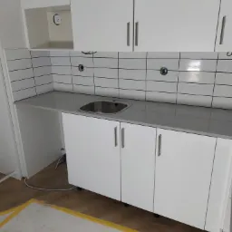 Nowoczesna, biała zabudowa kuchenna z szarą, kamienną płytą roboczą i prostokątnymi płytkami na ścianie, widoczne otwory na baterię i syfon, częściowo zabezpieczona podłoga taśmą malarską.