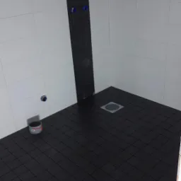 Niedokończona kabina prysznicowa z czarną, kwadratową mozaiką na podłodze i pionowym pasem na ścianie, białe płytki na ścianach, widoczny odpływ liniowy i poziomica.