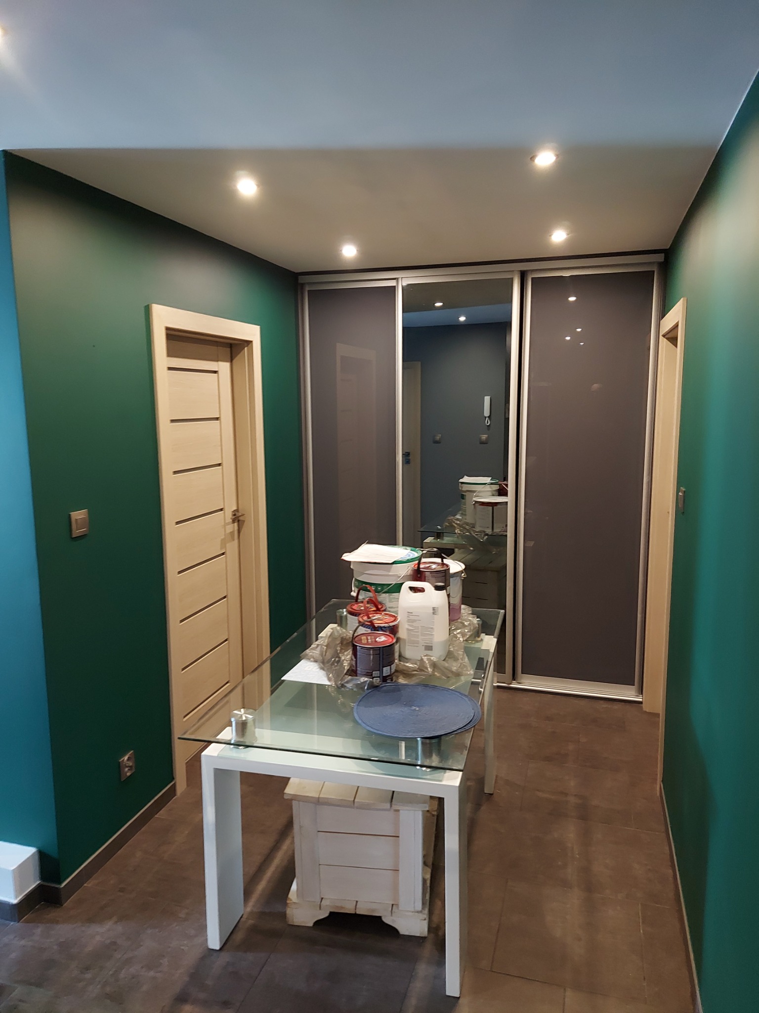 Wnętrze korytarza w trakcie prac wykończeniowych, ściany pomalowane na zielono, widoczne puszki z farbą na stole, zamontowane drzwi wewnętrzne z jasnego drewna oraz szafa przesuwna w zabudowie.