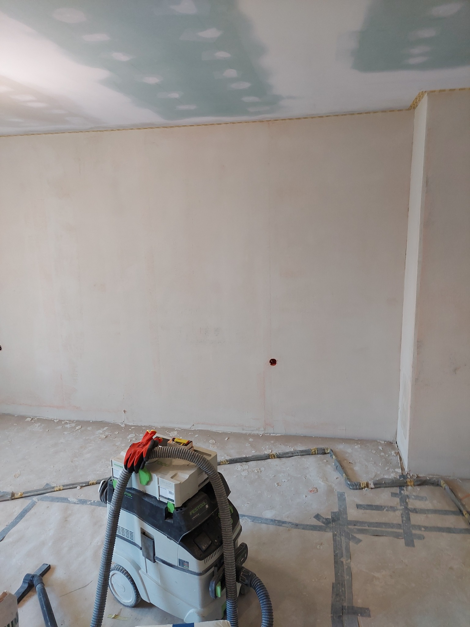 Wnętrze w trakcie prac wykończeniowych: ściany po nałożeniu gładzi, częściowo wykończony sufit, profesjonalny odkurzacz budowlany Festool z rękawicami roboczymi na górze, zabezpieczona podłoga.