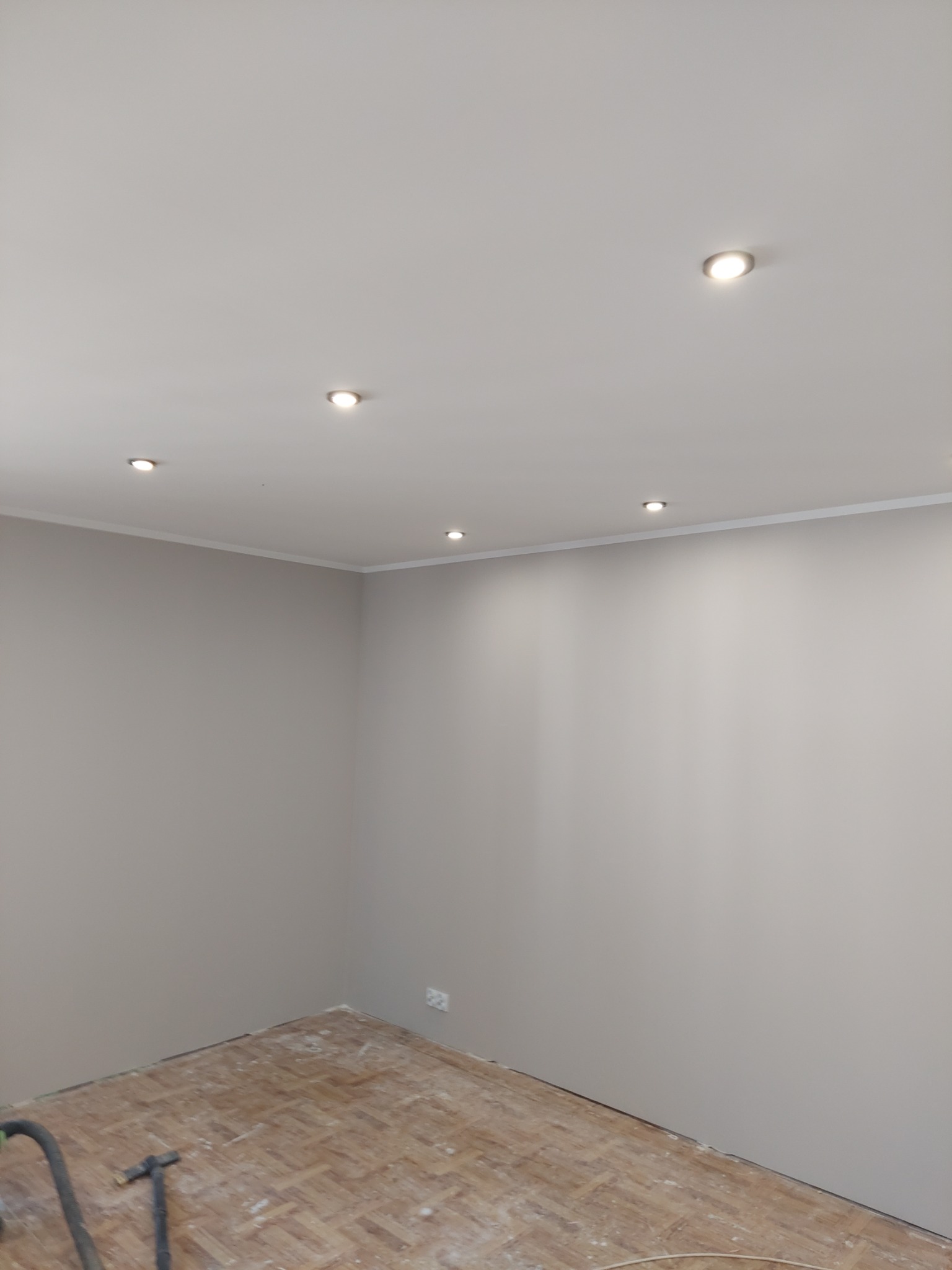 Pokój w trakcie odświeżania, ściany pomalowane na szaro, widoczne halogenowe oświetlenie sufitowe, stara drewniana podłoga przygotowana do dalszych prac, rura od odkurzacza przemysłowego przy...