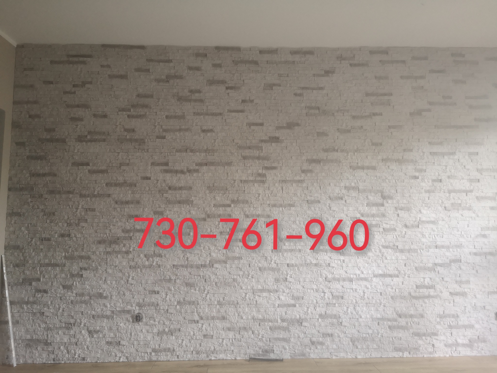 Ściana wykończona dekoracyjnym kamieniem w odcieniach szarości, widoczna w pomieszczeniu w Pabianicach. Na ścianie naniesiony czerwony numer telefonu.