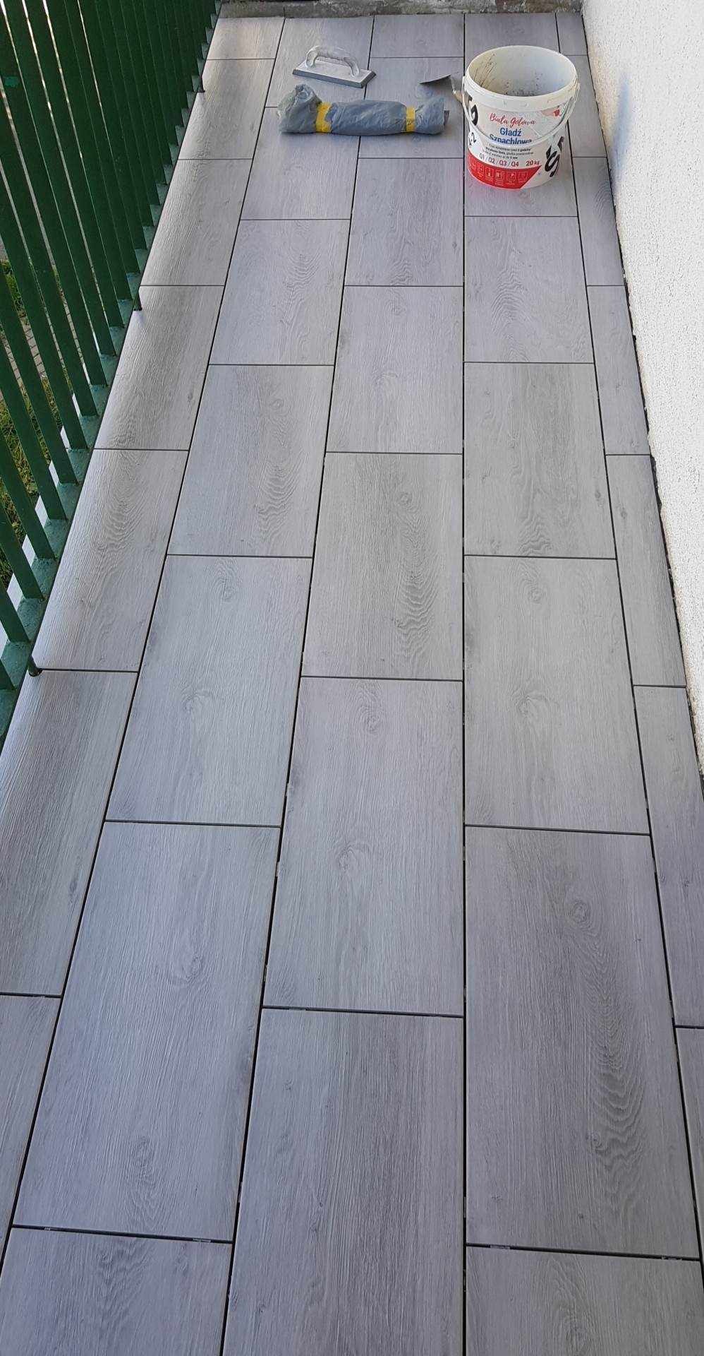 Szare płytki imitujące drewno na balkonie, widoczny fragment zielonej barierki, wiadro z zaprawą i narzędzia glazurnicze.