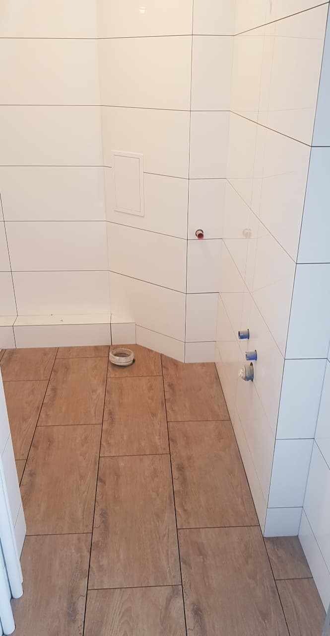 Wykończona łazienka z białymi kaflami ściennymi i panelami podłogowymi imitującymi drewno, widoczne przyłącza wodne i mała klapka rewizyjna.