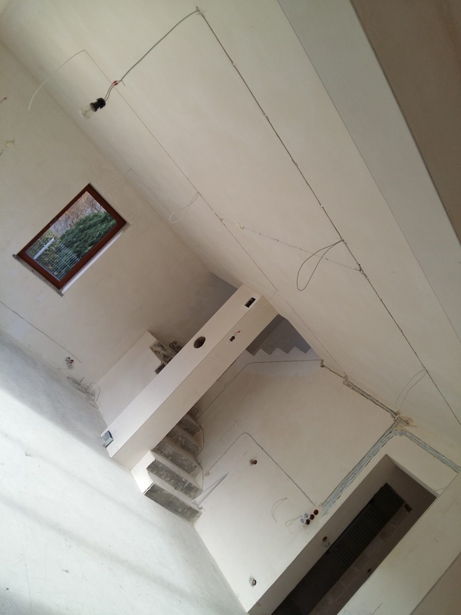 Poddasze w trakcie prac wykończeniowych: widoczne przewody elektryczne, okno dachowe, surowe schody i ściany po szpachlowaniu, przygotowane do dalszych prac.