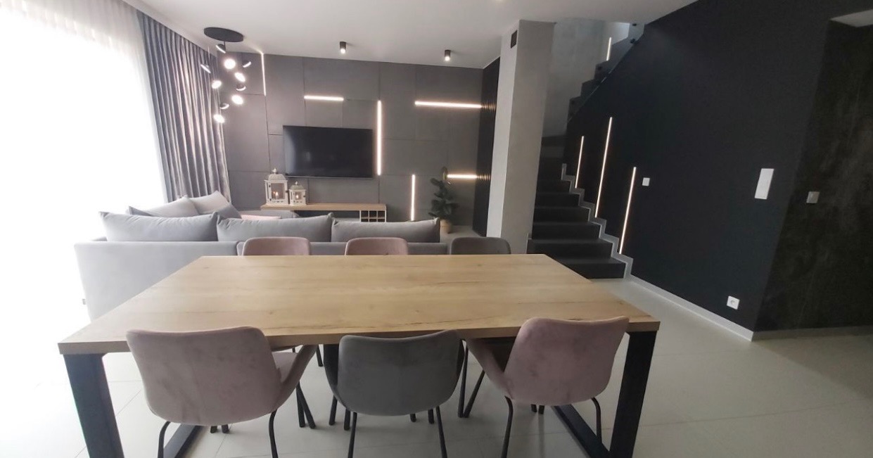 Nowoczesne wnętrze salonu z drewnianym stołem, welurowymi krzesłami w odcieniach różu i szarości, szarą ścianą z podświetleniem LED i schodami w tle.