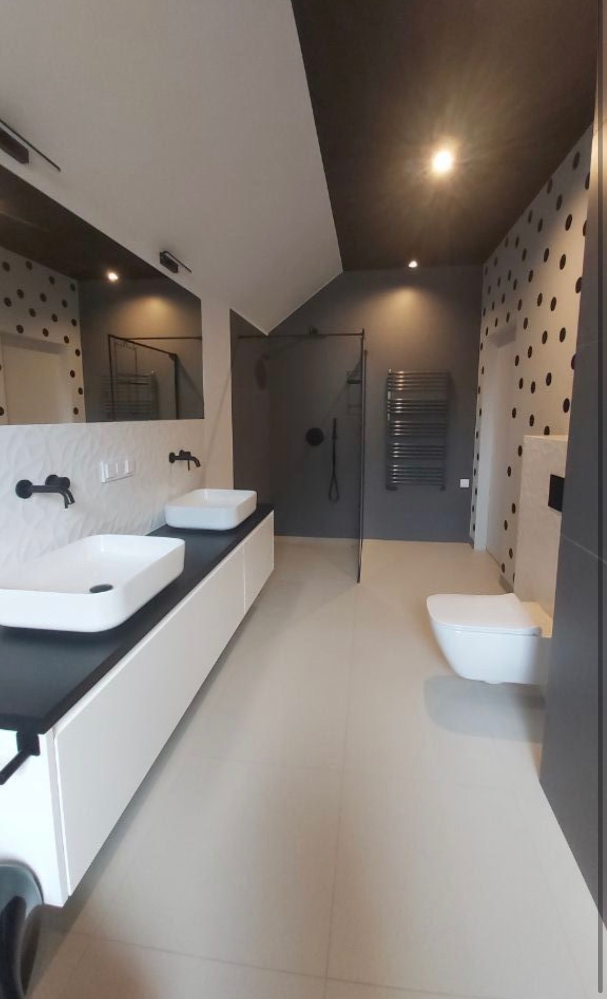 Nowoczesna łazienka z dwoma umywalkami nablatowymi na białej szafce z czarnym blatem, czarną kabiną prysznicową typu walk-in i grzejnikiem drabinkowym na szarej ścianie, oraz toaletą i ścianą...