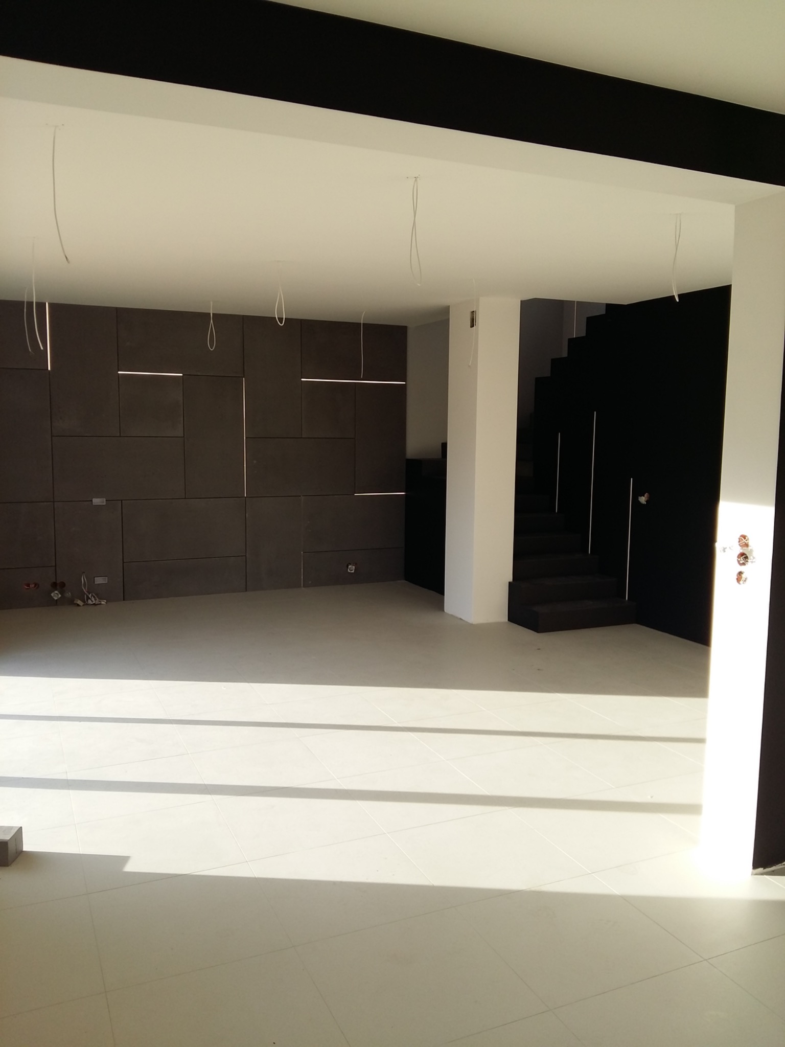 Jasne, przestronne wnętrze w trakcie wykańczania: ściana z paneli z podświetleniem, biała podłoga z płytek, czarne schody i sufity, widoczne kable oświetleniowe.