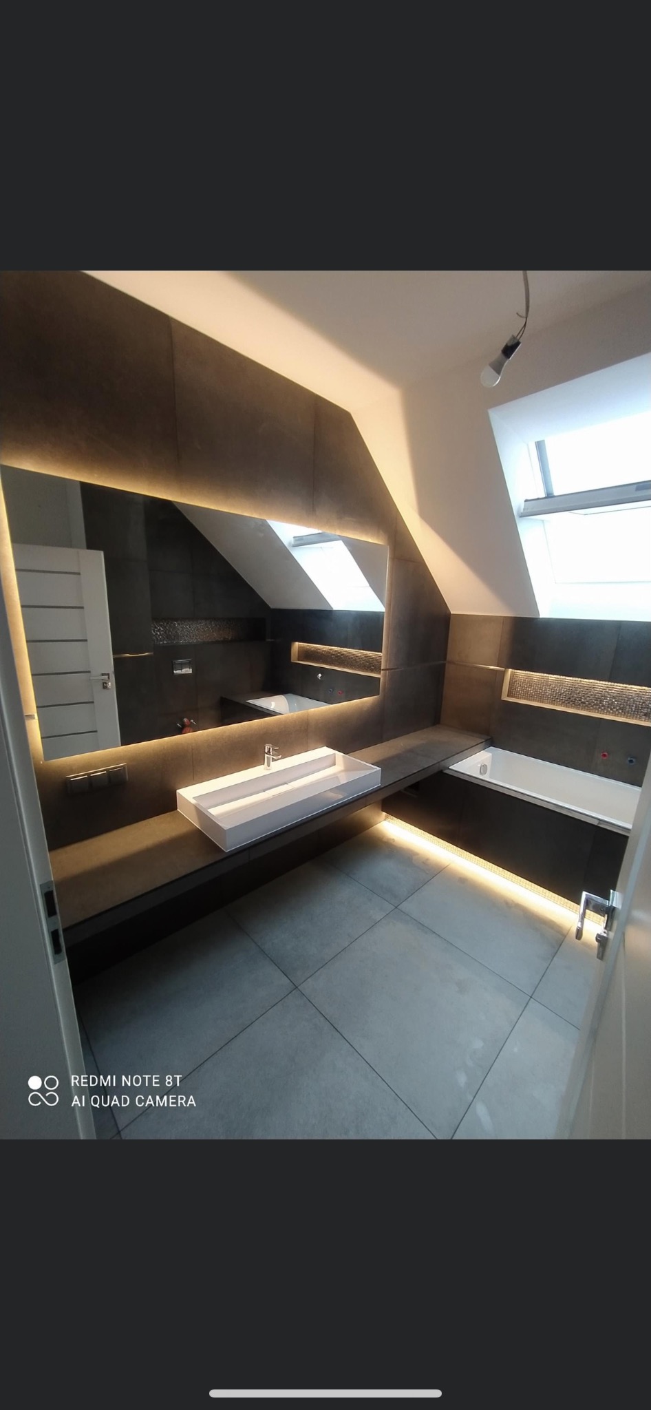 Nowoczesna łazienka na poddaszu z szarą glazurą, podświetlanym lustrem i umywalką w kształcie koryta, widok z perspektywy wejścia.