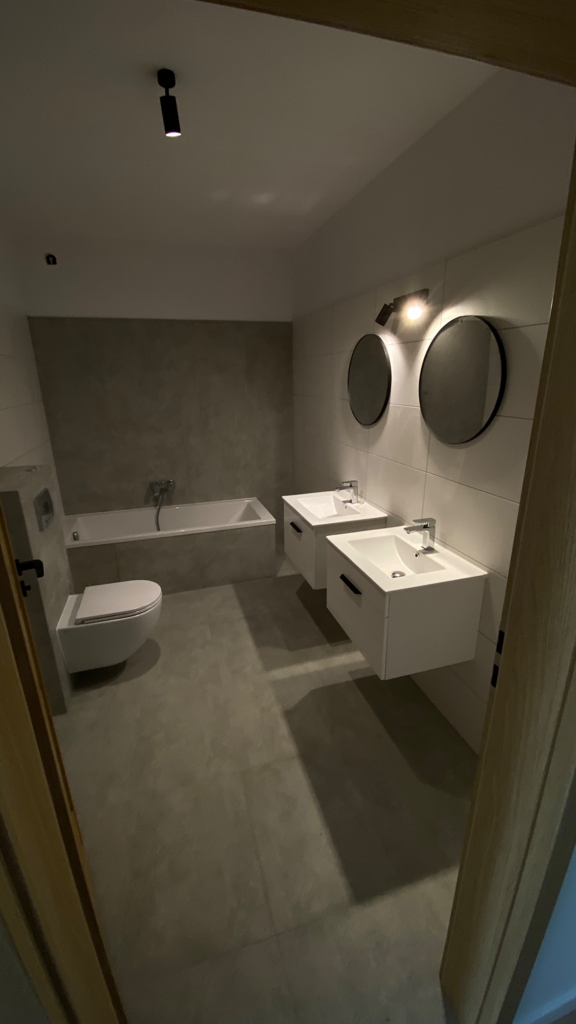 Nowoczesna łazienka z betonową ścianą, wanną, podwieszaną toaletą i dwoma umywalkami z szafkami, oświetlona punktowym światłem i okrągłymi lustrami.