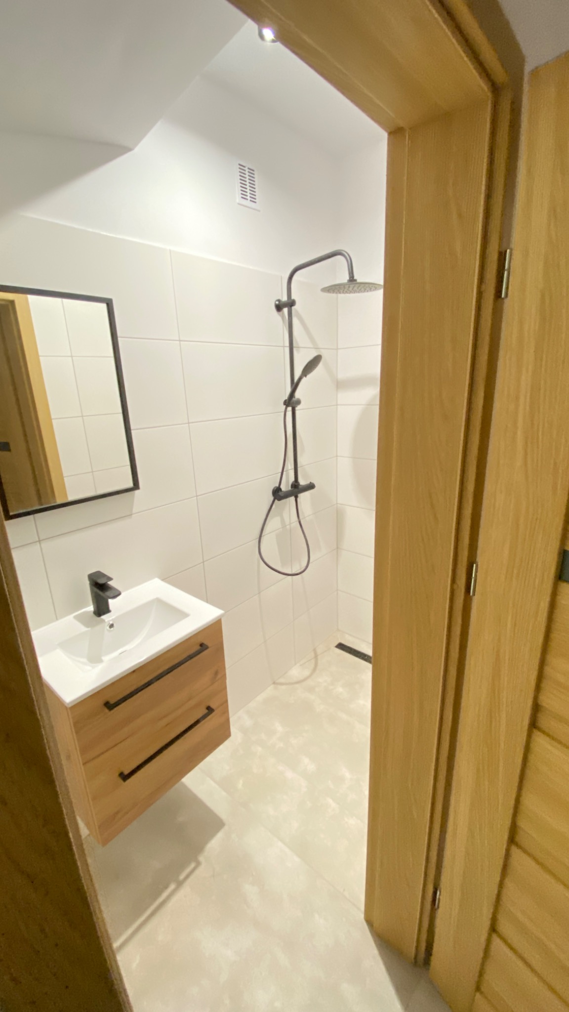 Nowoczesna łazienka z kabiną prysznicową typu walk-in, umywalką z szafką z drewna i czarną armaturą, widok zza drewnianej ościeżnicy.