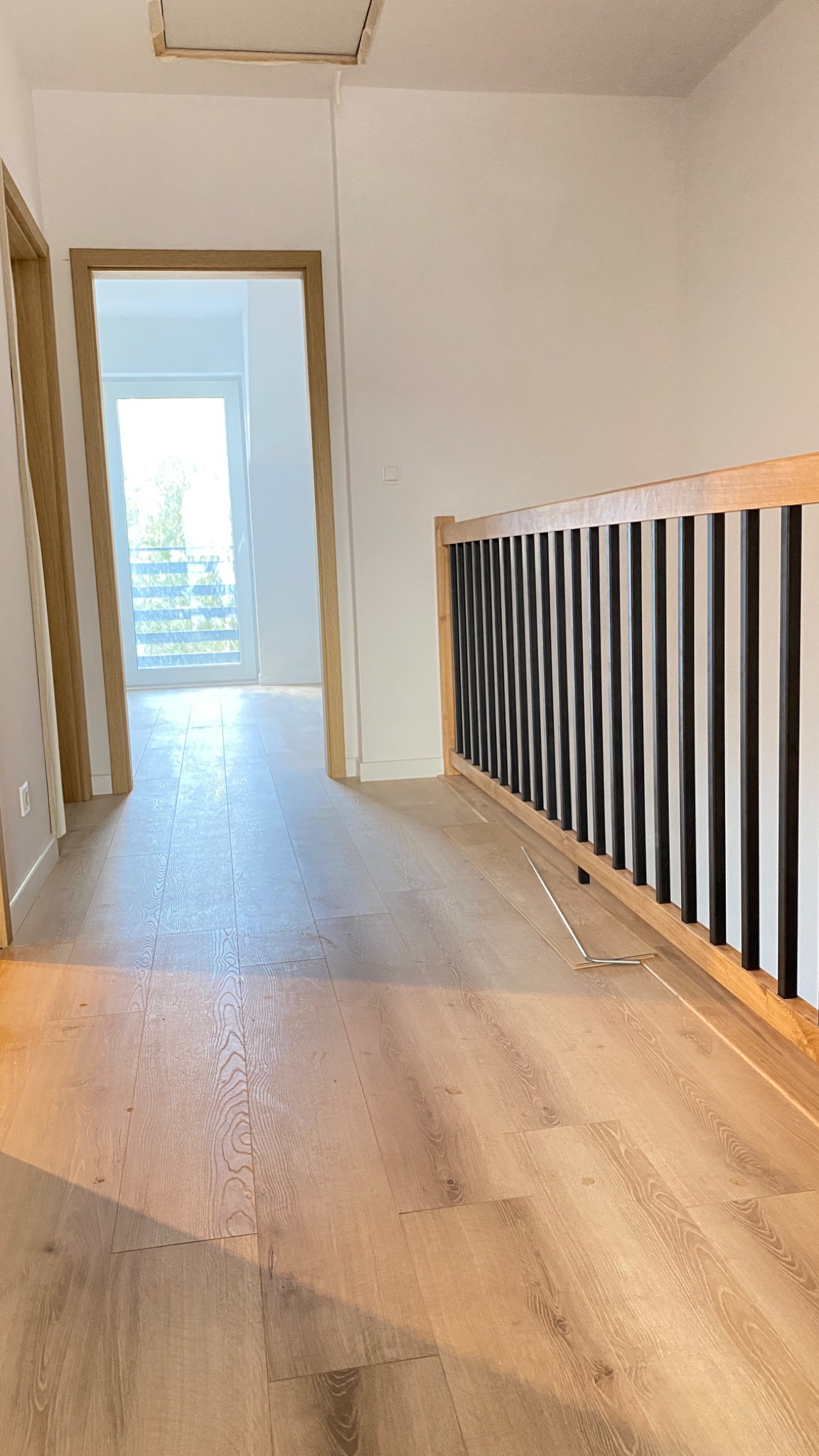 Jasny korytarz z ułożonym parkietem w odcieniach beżu, widoczne fragmenty niezamontowanych desek parkietowych przy balustradzie schodów z czarnymi szczeblami i drewnianą ramą, w tle otwarte drzwi...