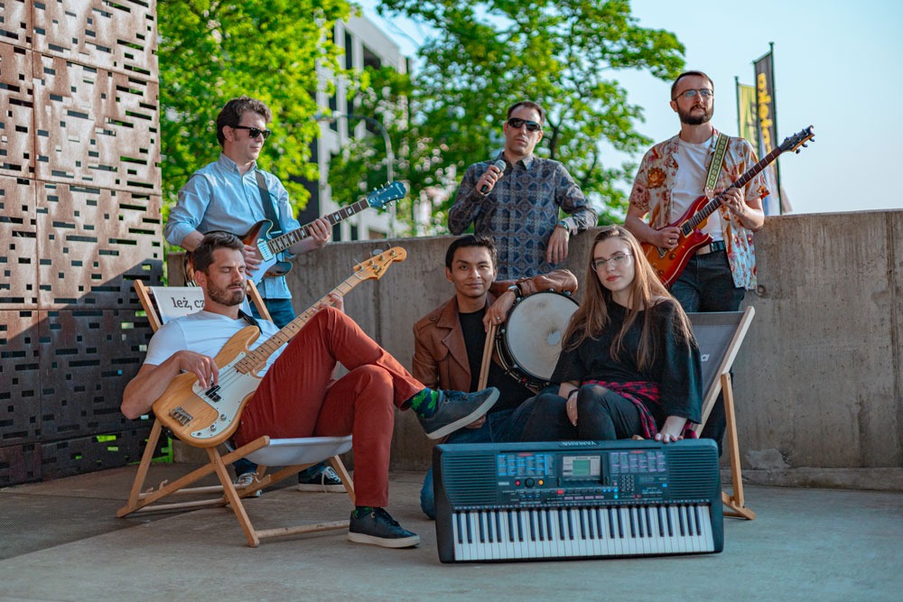 Zespół muzyczny pozuje z instrumentami: gitary, bas, keyboard, perkusja, mikrofon na tle miejskiej architektury i zieleni.