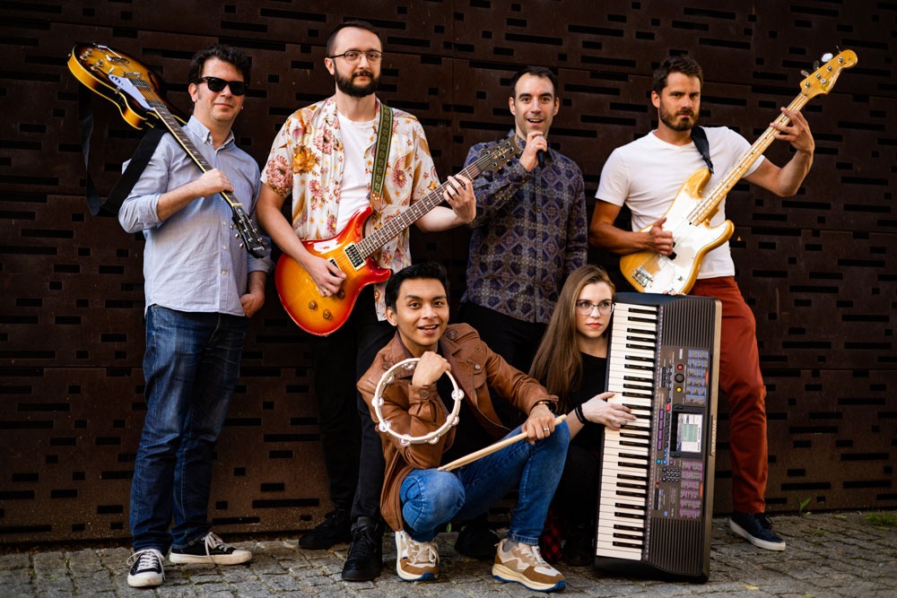 Zespół muzyczny w składzie sześcioosobowym prezentuje się z instrumentami: gitara elektryczna, gitara basowa, keyboard, tamburyn i mikrofon na tle metalowej ściany.