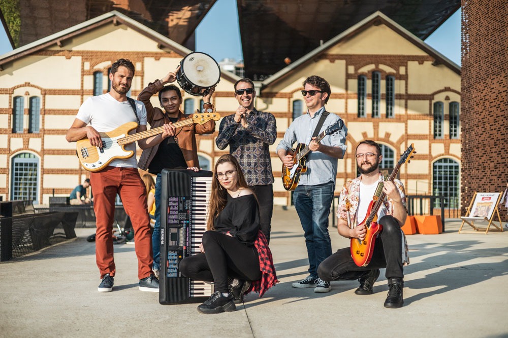 Zespół muzyczny w plenerze, sześciu muzyków z instrumentami: gitary, bas, keyboard, perkusja, pozuje na tle ceglanych budynków.