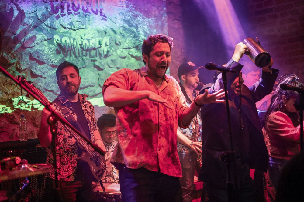 Zespół muzyczny grający koncert na scenie z oświetleniem scenicznym, wokalista w czerwonej koszuli śpiewa z ekspresją, basista po lewej stronie, perkusista w tle, mężczyzna w garniturze trzyma...