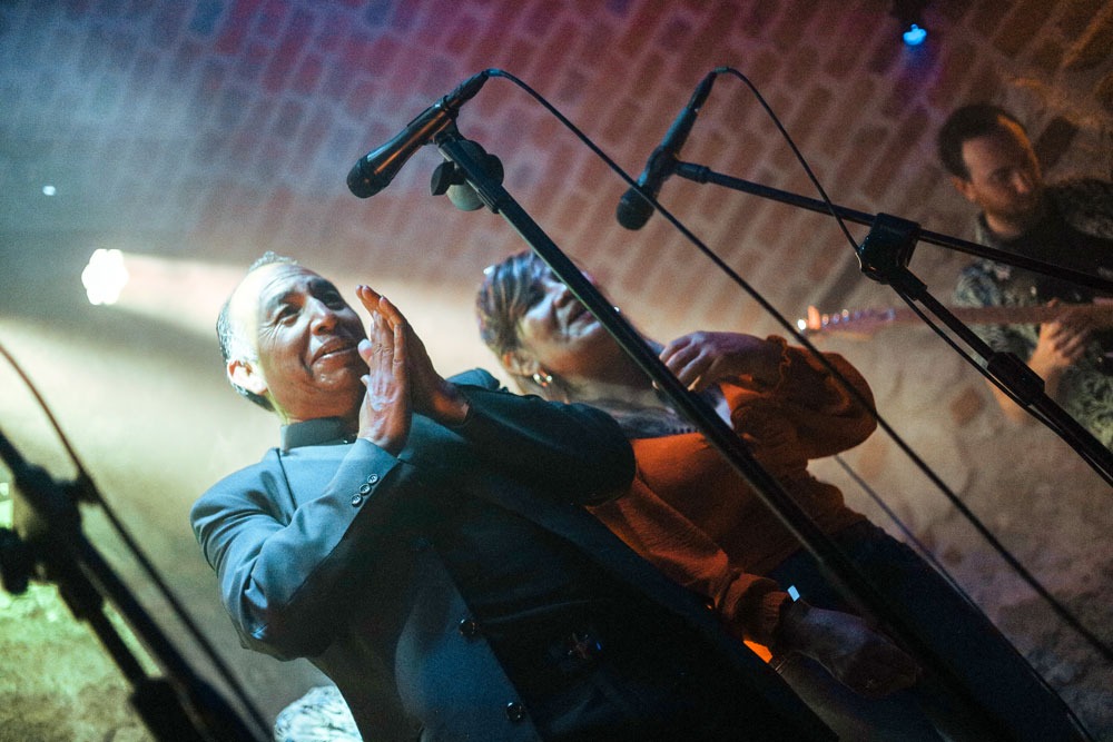 Zespół muzyczny podczas występu na scenie, mężczyzna w garniturze klaszcze, w tle kobieta w pomarańczowej bluzce oraz gitarzysta