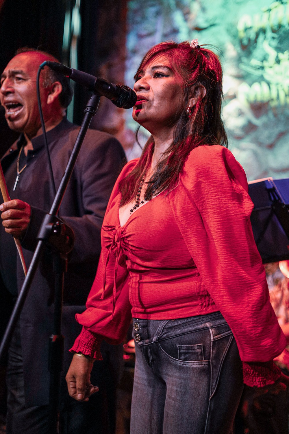 Kobieta w czerwonej bluzce śpiewa do mikrofonu podczas występu na scenie z perkusistą w tle.