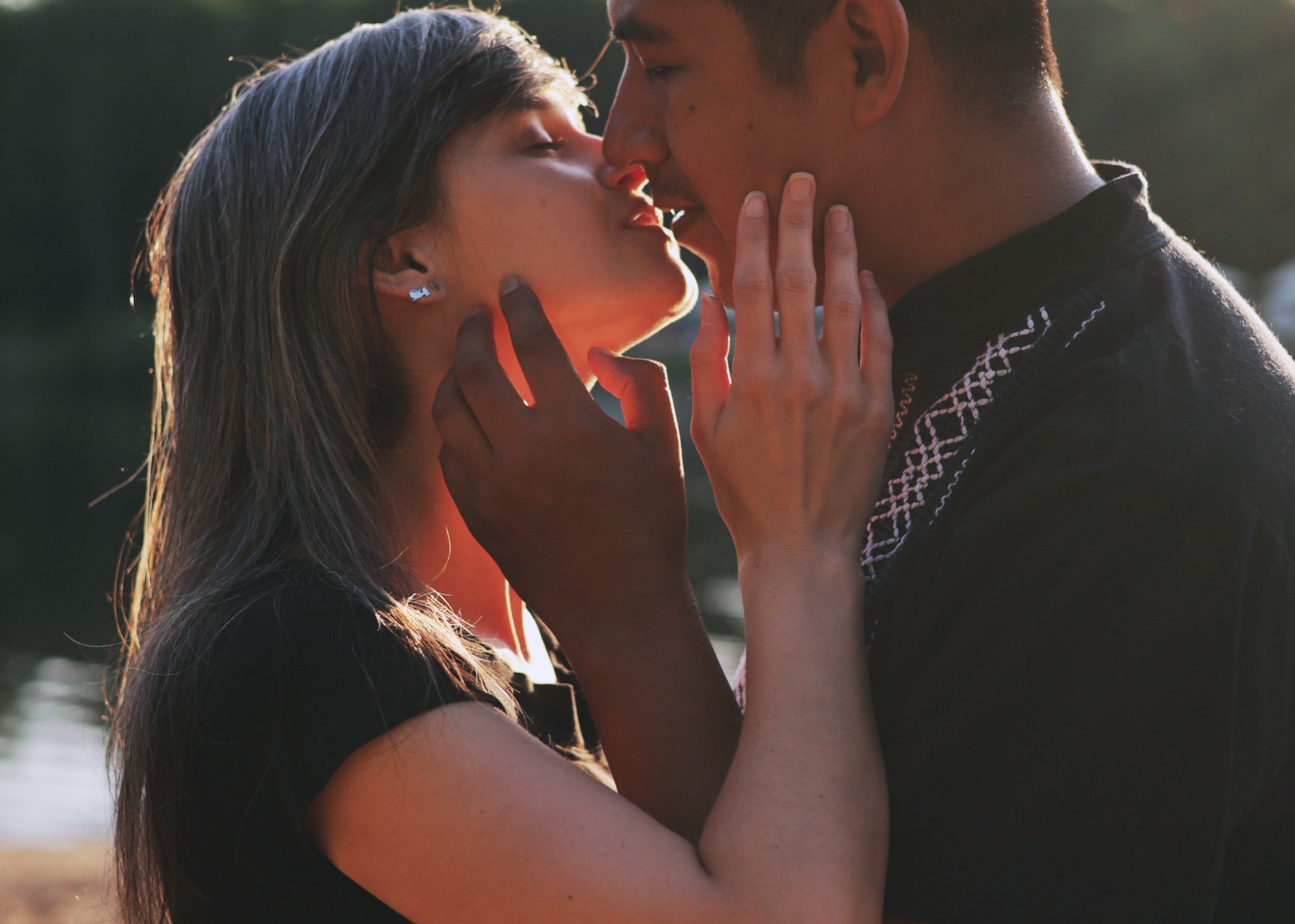 Para całująca się w ciepłym świetle, dłoń kobiety spoczywa na policzku mężczyzny, widoczny detal koszuli z haftem.