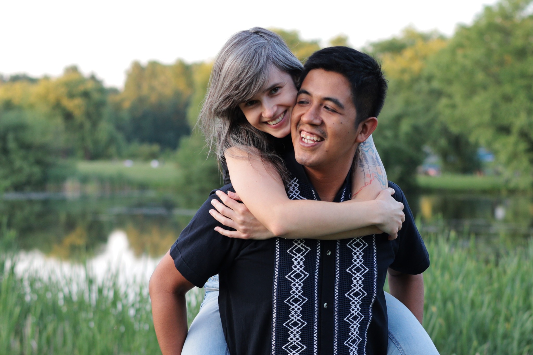 Uśmiechnięta kobieta z siwymi włosami obejmuje szyję mężczyzny z tatuażem na ramieniu, siedząc na jego plecach na tle jeziora i zieleni.