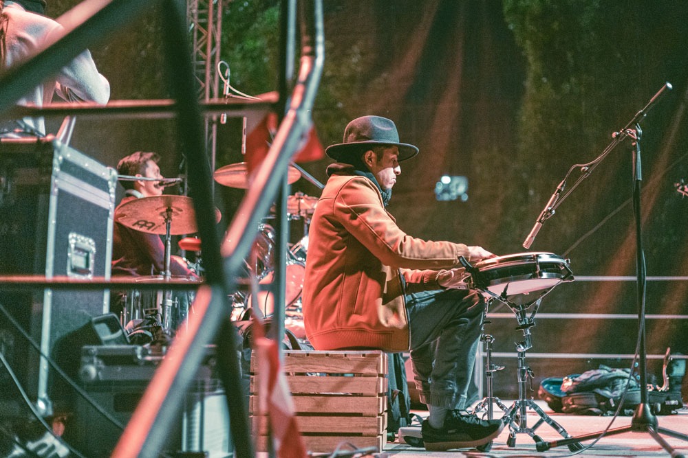 Muzyk w kapeluszu gra na instrumencie perkusyjnym podczas występu na scenie, w tle widoczny perkusista i sprzęt nagłośnieniowy.