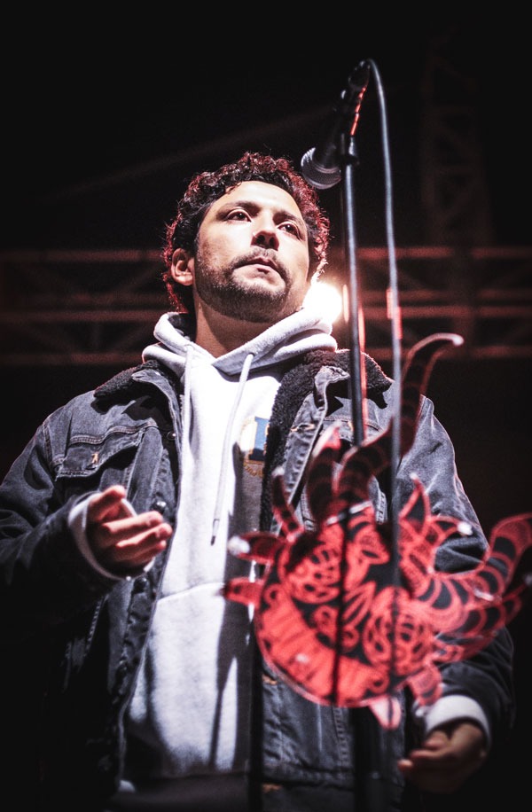 Mężczyzna z kręconymi włosami w dżinsowej kurtce i bluzie z kapturem, stojący przy mikrofonie na scenie, z dekoracją w kształcie słońca w tle.