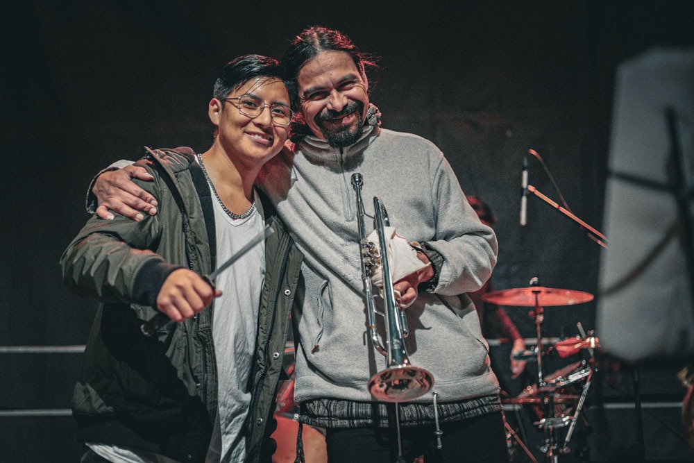 Dwóch uśmiechniętych mężczyzn pozuje z puzonem na scenie muzycznej, w tle widoczne perkusja i mikrofony.