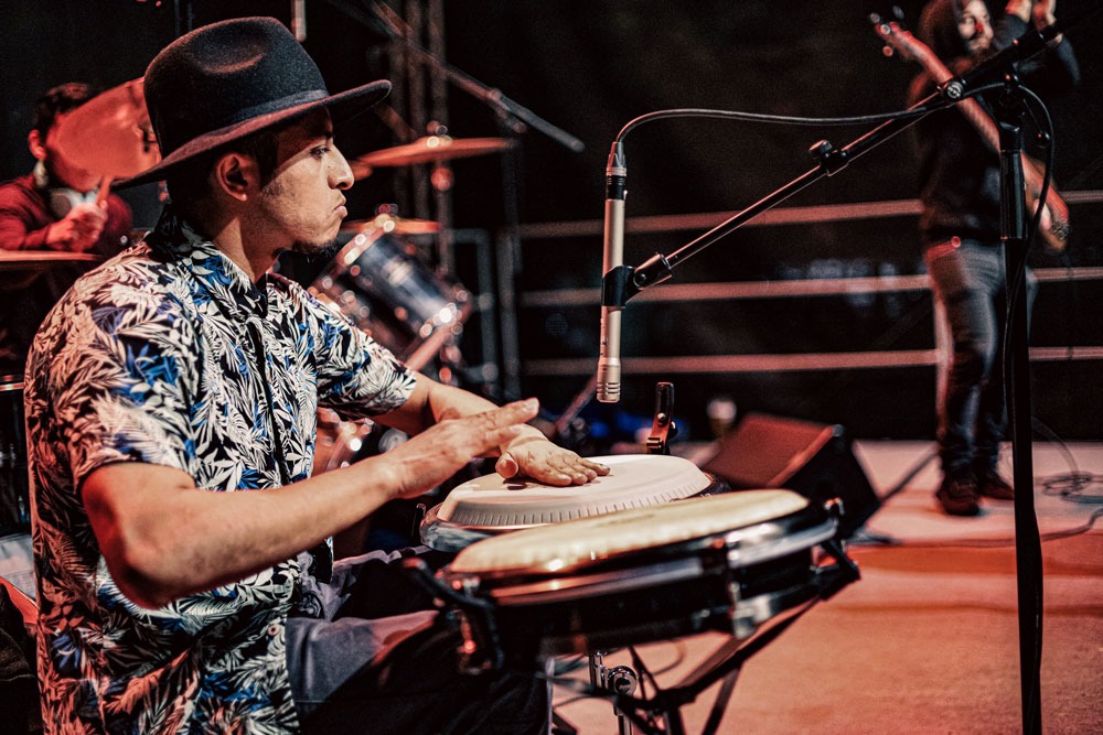 Muzyk w kapeluszu grający na bębnach conga podczas występu na scenie, z mikrofonem i innymi muzykami w tle.