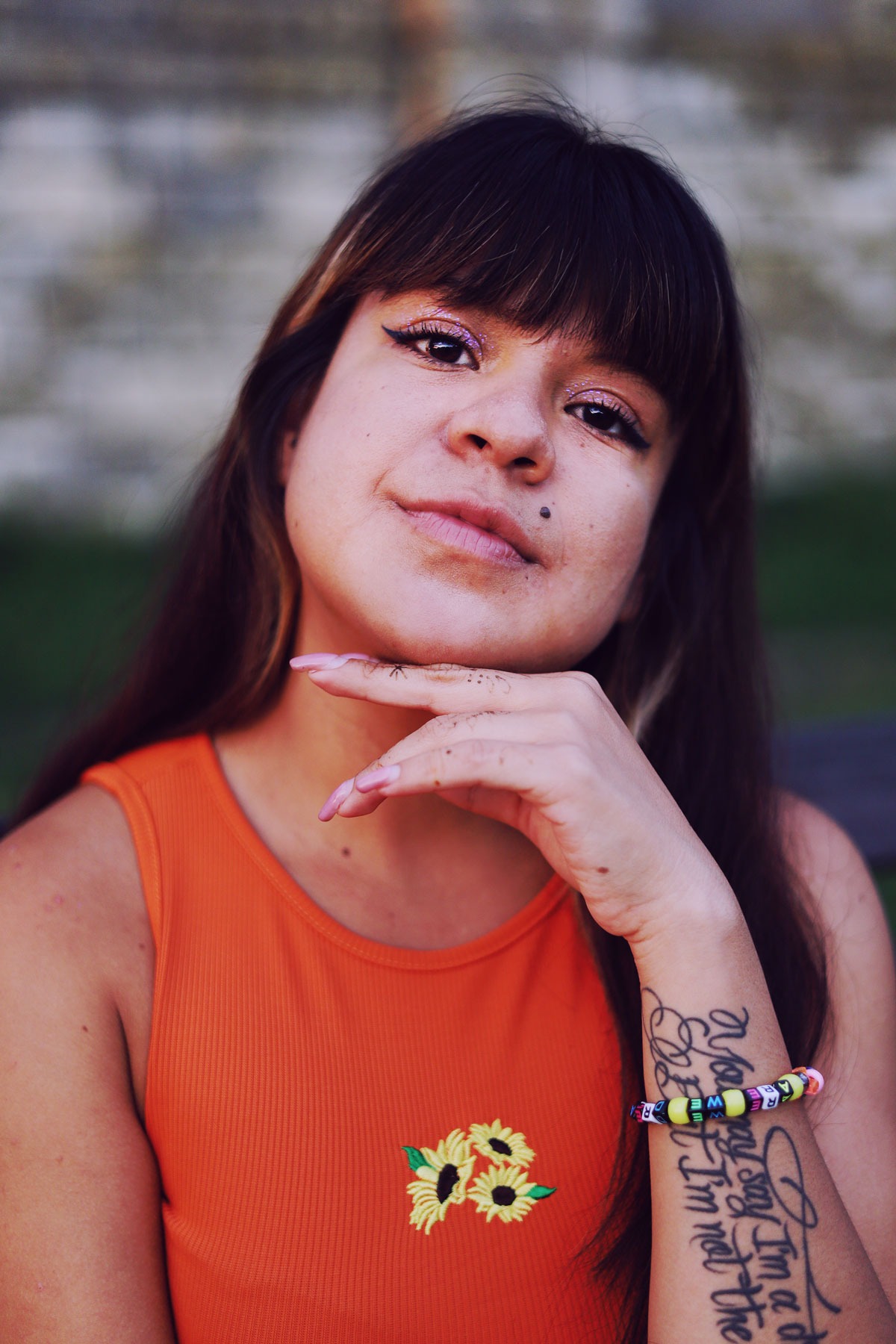 Portret kobiety z ciemnymi włosami i grzywką, w pomarańczowej bluzce z haftowanymi słonecznikami, z brokatowym makijażem oczu i tatuażami na rękach, pozującej z dłonią pod brodą.