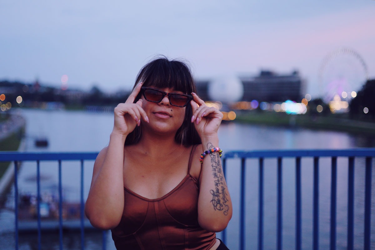 Portret kobiety w okularach przeciwsłonecznych na tle rzeki i wesołego miasteczka w Krakowie, widoczny tatuaż na ramieniu.