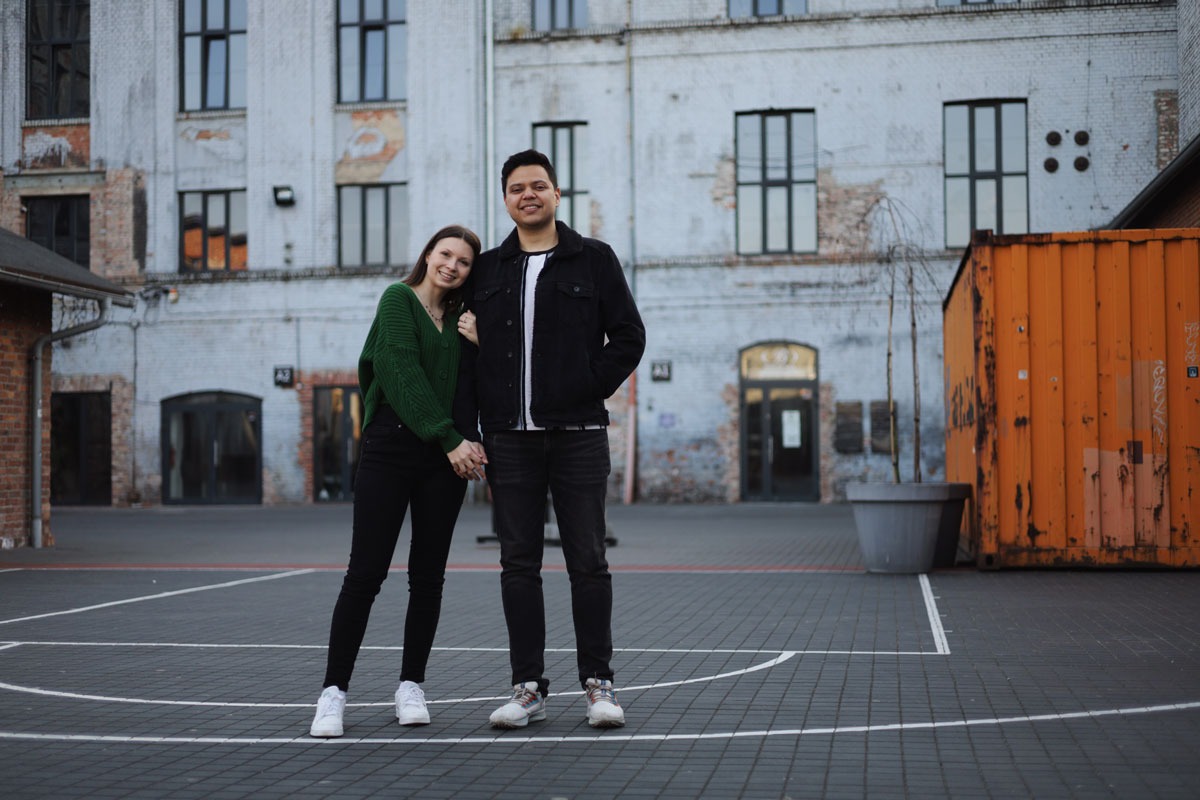 Uśmiechnięta para trzymająca się za ręce na tle miejskiego dziedzińca z ceglanym budynkiem i pomarańczowym kontenerem.