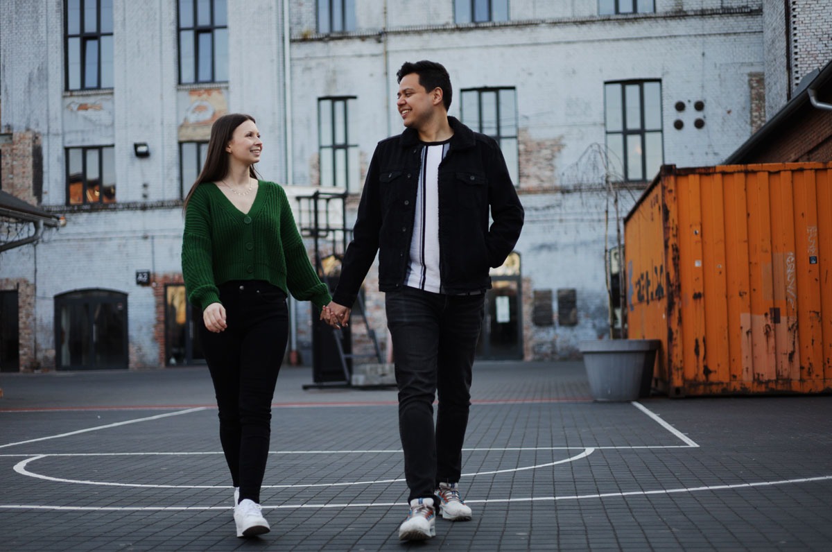 Para trzymająca się za ręce spaceruje po placu z budynkami w tle, kobieta w zielonym swetrze i mężczyzna w czarnej kurtce uśmiechają się do siebie.
