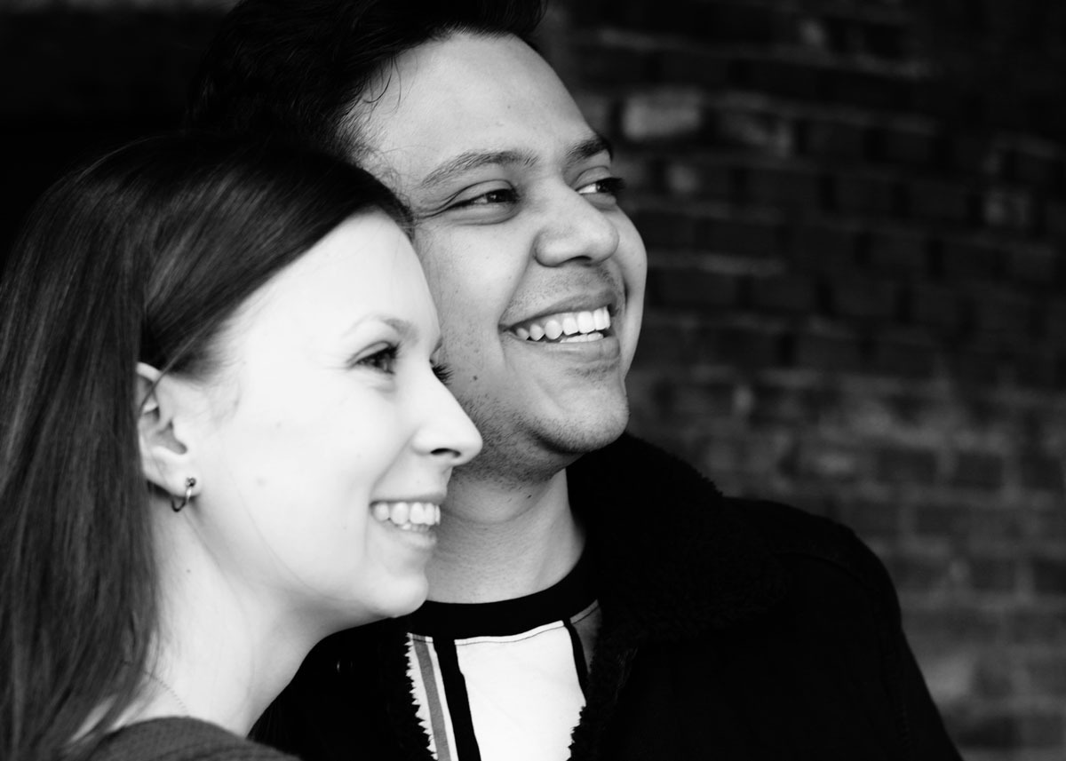 Czarno-białe zbliżenie uśmiechniętej pary, kobieta z lewej strony, mężczyzna obok niej, oboje patrzą w prawo, ceglana ściana w tle.