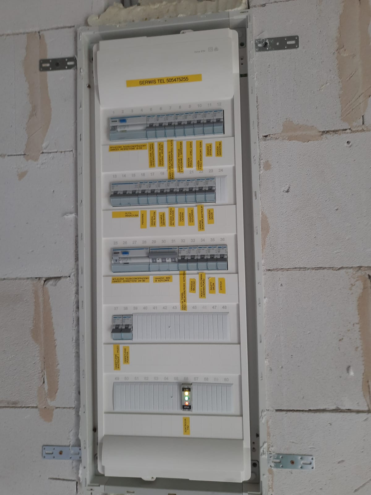 Rozdzielnica elektryczna z widocznymi bezpiecznikami, opisami obwodów i kontrolkami LED, zamontowana na surowej ścianie z betonu komórkowego, z żółtą naklejką z numerem telefonu serwisu.