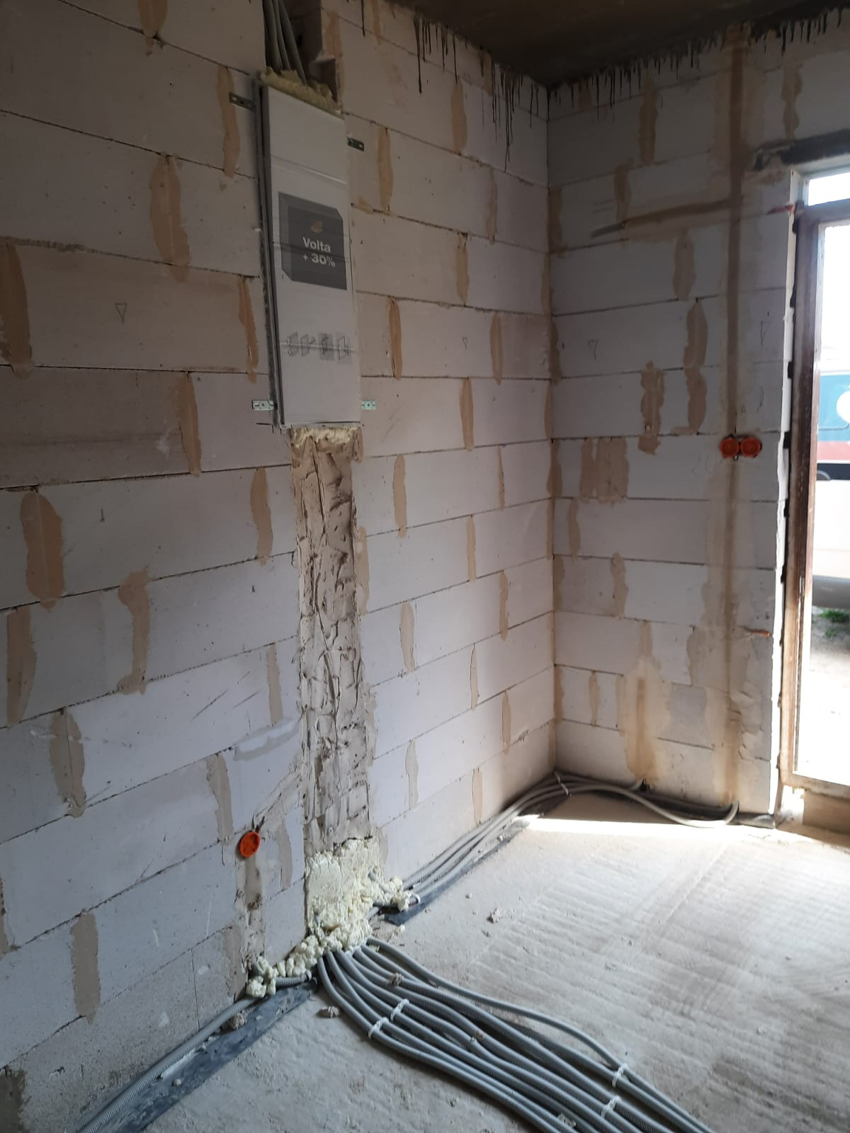 Wnętrze w stanie surowym z widoczną rozdzielnicą elektryczną Volta +30% na ścianie z betonu komórkowego, kable elektryczne wychodzące ze ściany zabezpieczone pianką montażową, oraz pomarańczowa...