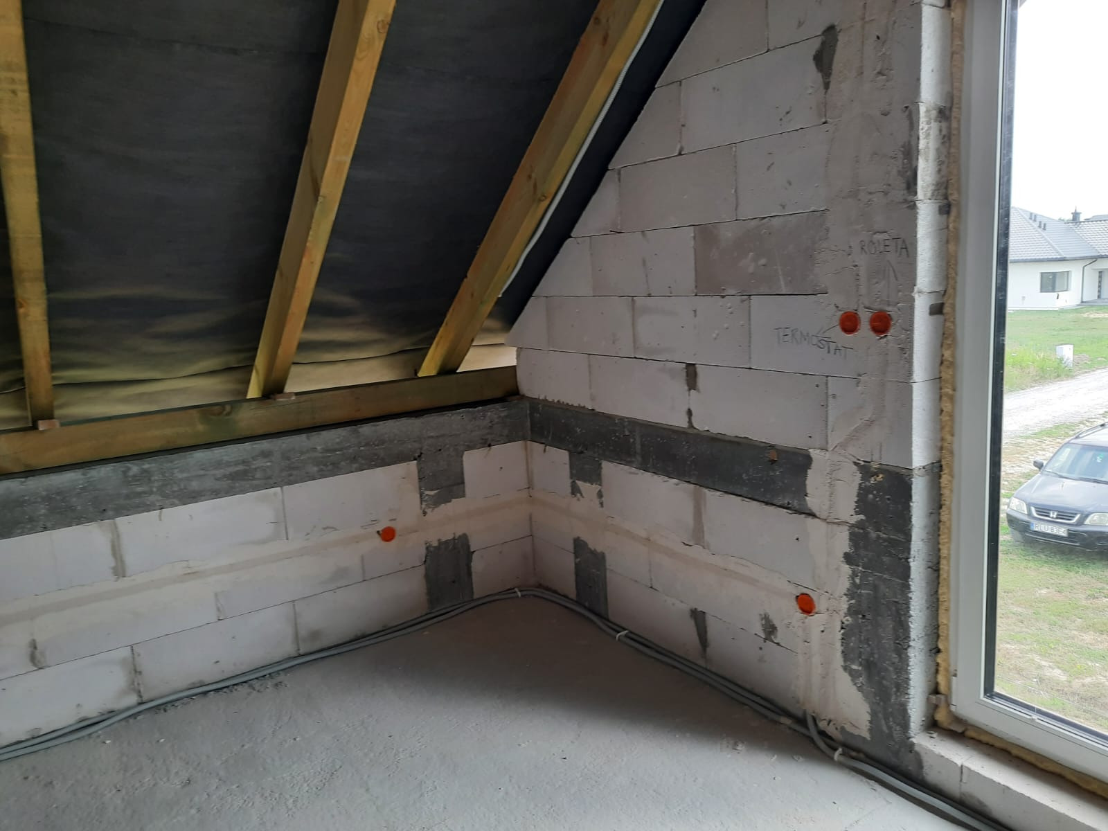 Surowy stan poddasza w trakcie prac: widoczne przewody elektryczne w szarych rurkach ułożone wzdłuż ściany z białych bloczków, otwory pod gniazdka, drewniana konstrukcja dachu z czarną membraną...