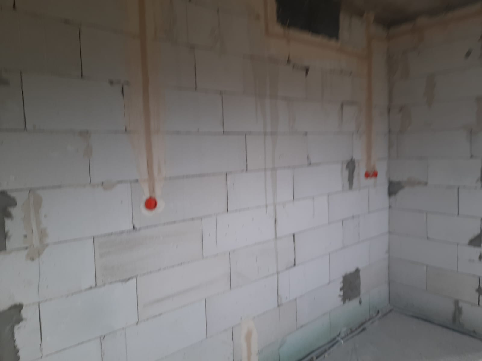 Surowa ściana z betonu komórkowego z pionowymi bruzdami wypełnionymi gipsem, w których poprowadzone są przewody elektryczne zakończone czerwonymi puszkami pod gniazdka i włączniki.