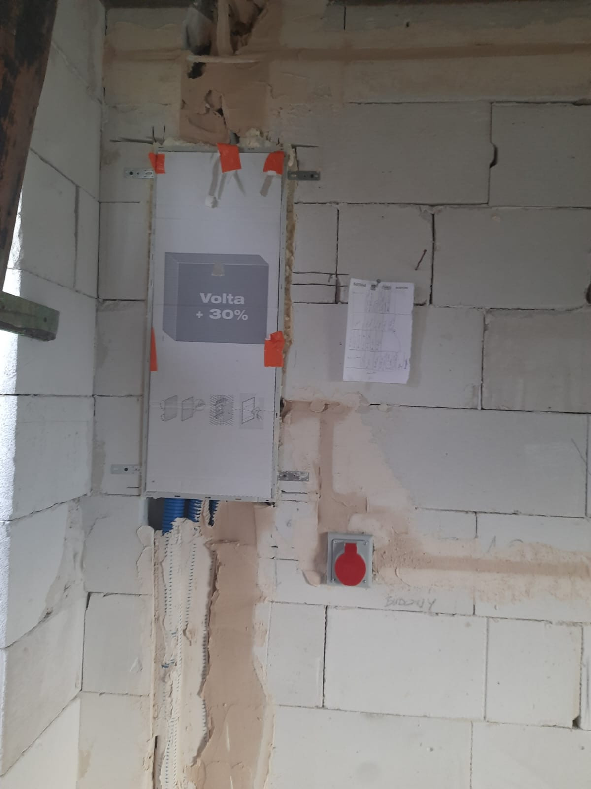 Wnętrze w trakcie remontu z zamontowaną, częściowo zakrytą skrzynką elektryczną Volta +30% na ścianie z bloczków, widoczne niebieskie peszle i wystające przewody, obok schemat instalacji i gniazdo...