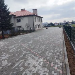 Nowo ułożona, szara kostka brukowa z czerwonymi akcentami na dużej powierzchni przed budynkiem, zakończona krawężnikiem i ogrodzeniem z siatki.