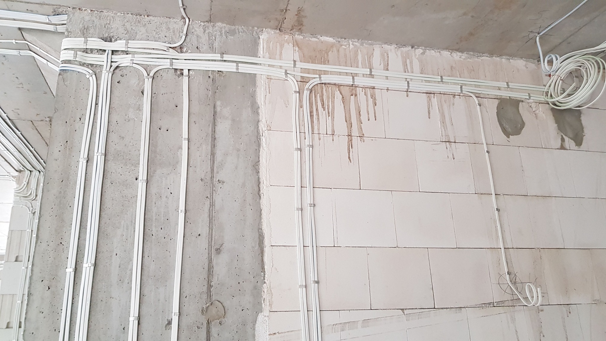 Białe przewody elektryczne prowadzone po surowych ścianach z betonu i bloczków, zabezpieczone uchwytami, widoczne ślady po zalaniu na ścianie z bloczków.