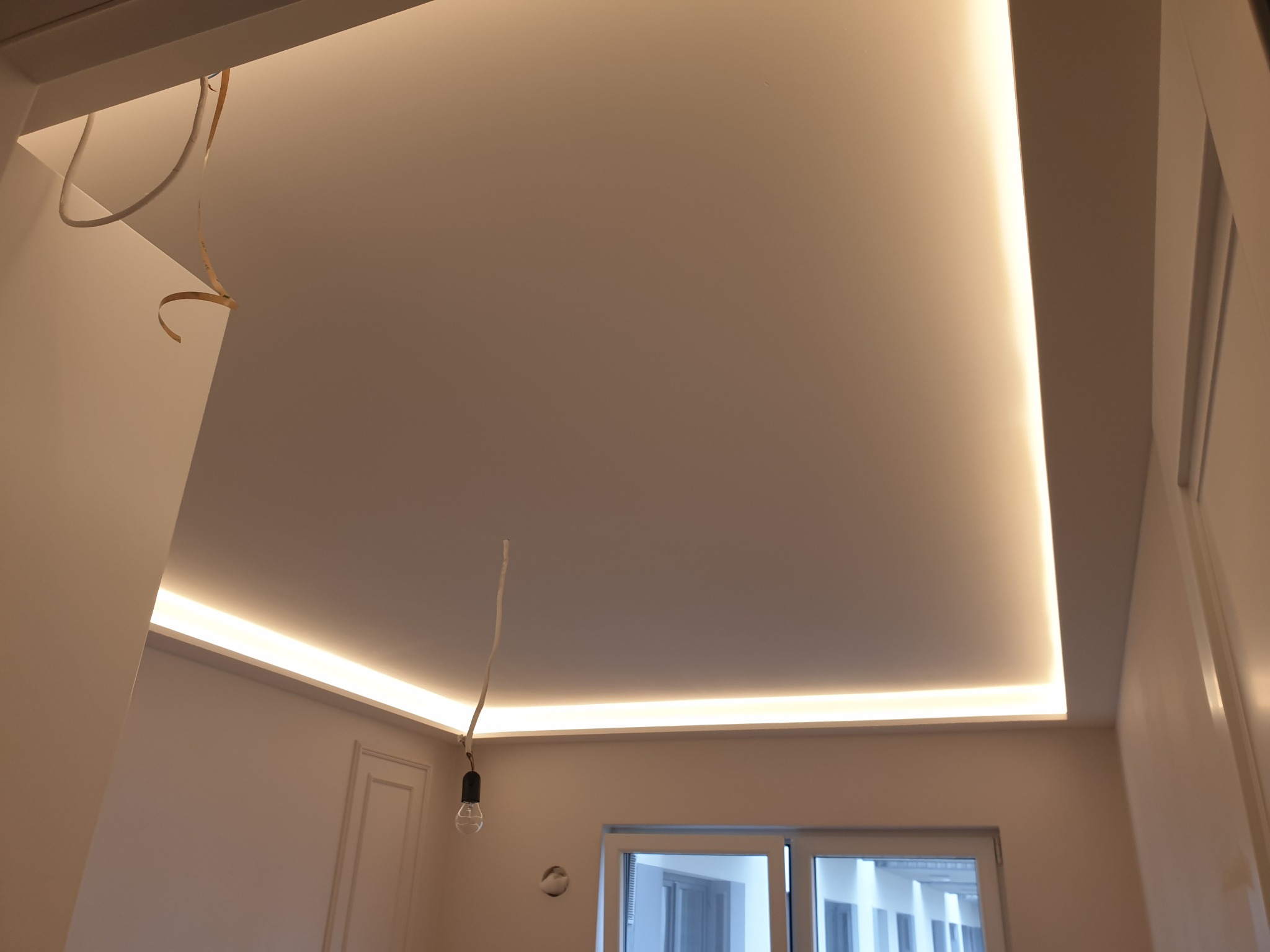 Podwieszany sufit z oświetleniem LED wzdłuż krawędzi, widok z dołu, w trakcie prac wykończeniowych w Warszawie. Wisząca żarówka na kablu i wystające przewody sugerują etap instalacji.