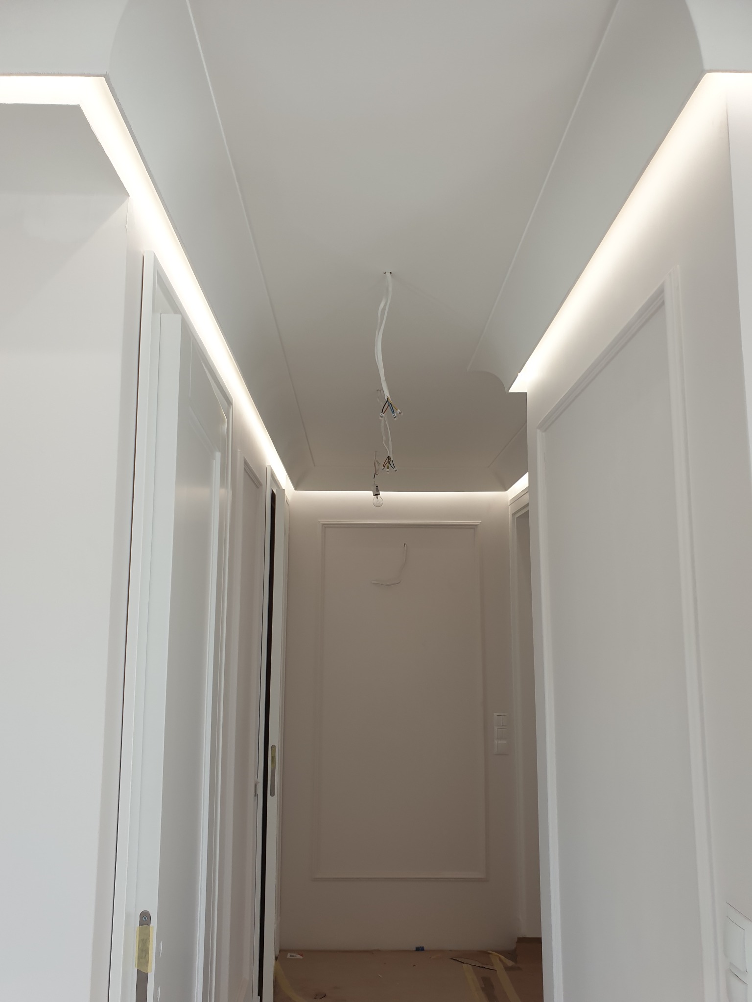 Korytarz w trakcie remontu z zamontowanym oświetleniem LED w listwach sufitowych i wiszącymi przewodami elektrycznymi w centralnej części sufitu.