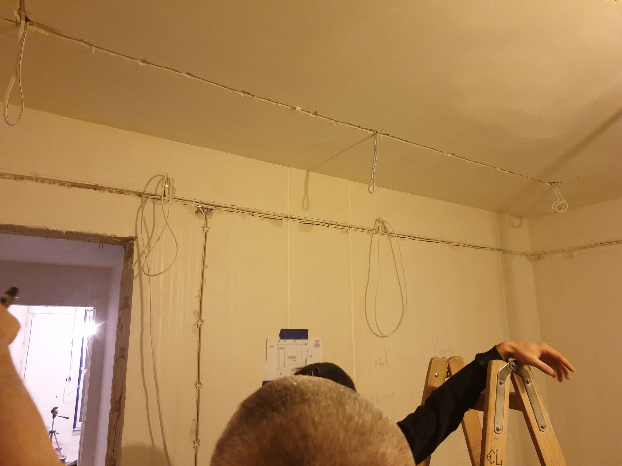 Wnętrze pokoju w trakcie prac elektrycznych: kable prowadzone po ścianach i suficie, widoczne mocowania i pętle kabli, osoba na drabinie, na ścianie plan instalacji zabezpieczony taśmą.